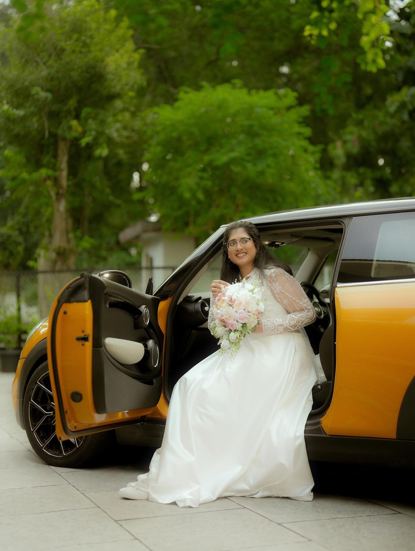 A bride seated beside an orange car with her bouquet